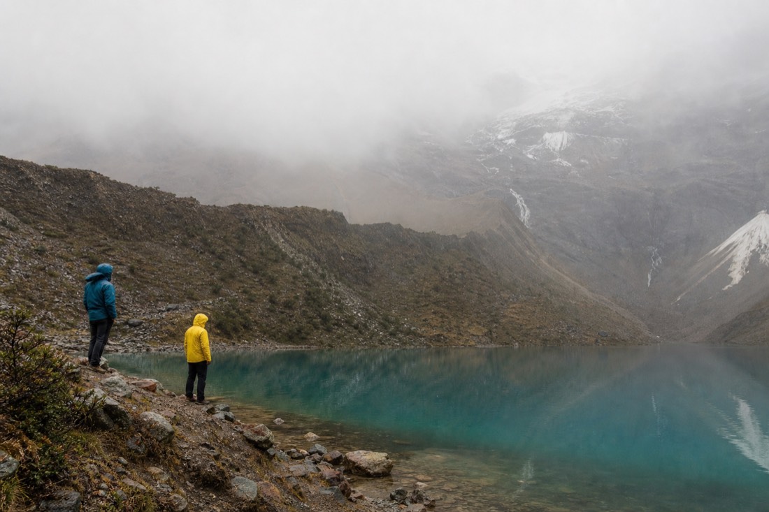 Hikers in rain jackets standing at the misty grey shoreline of Humantay Lake
