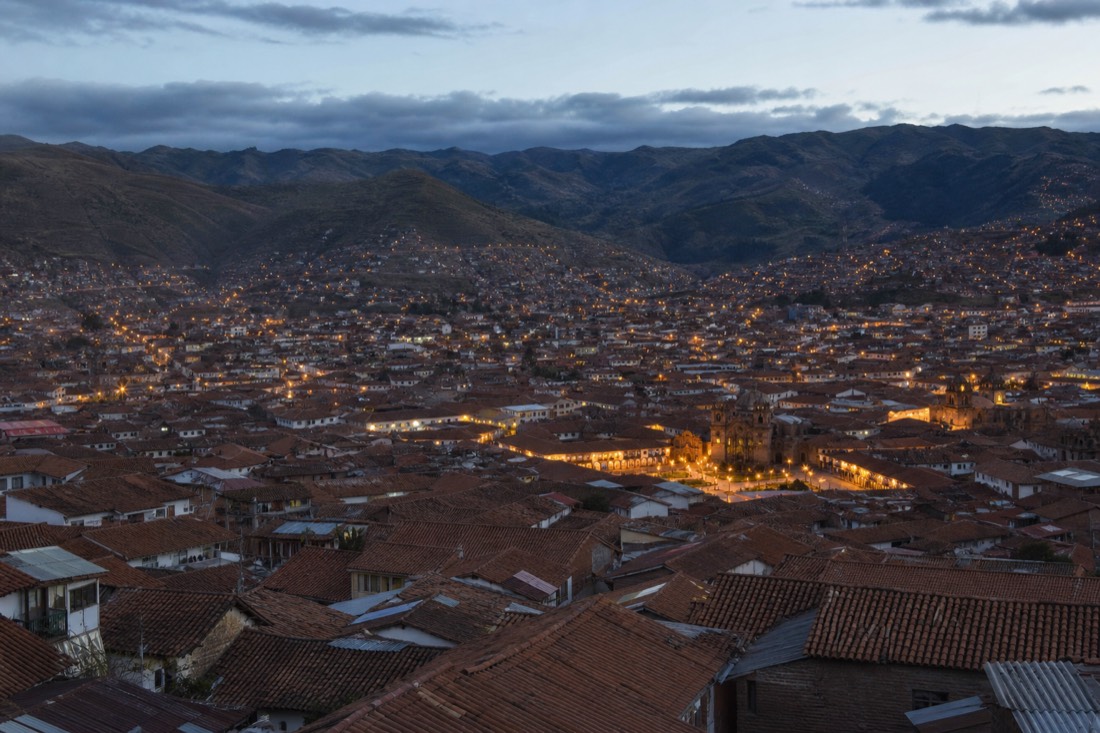 Terracotta rooftops of Cusco at dusk from a high vantage, warm lights coming on across the valley and dark hills behind