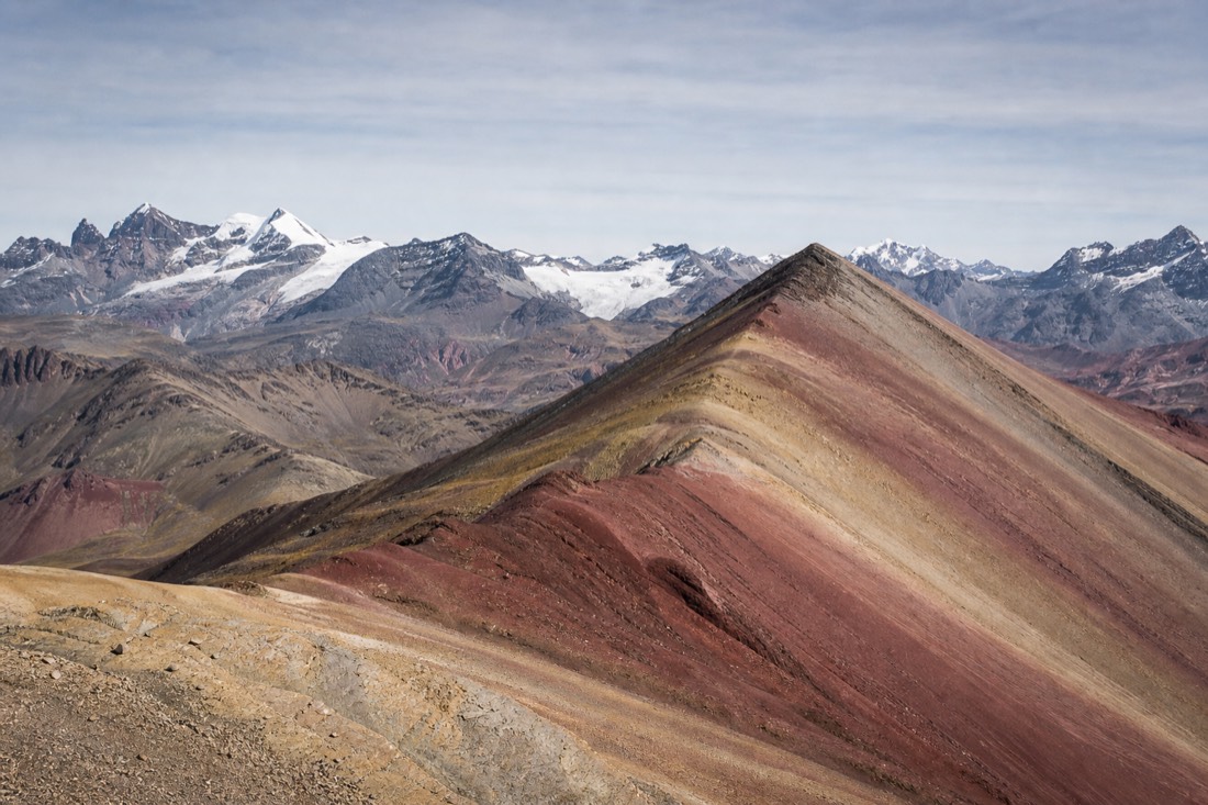 Wind-scoured high Andean ridge of red and ochre mineral bands under a pale cold sky with distant glaciers and no people