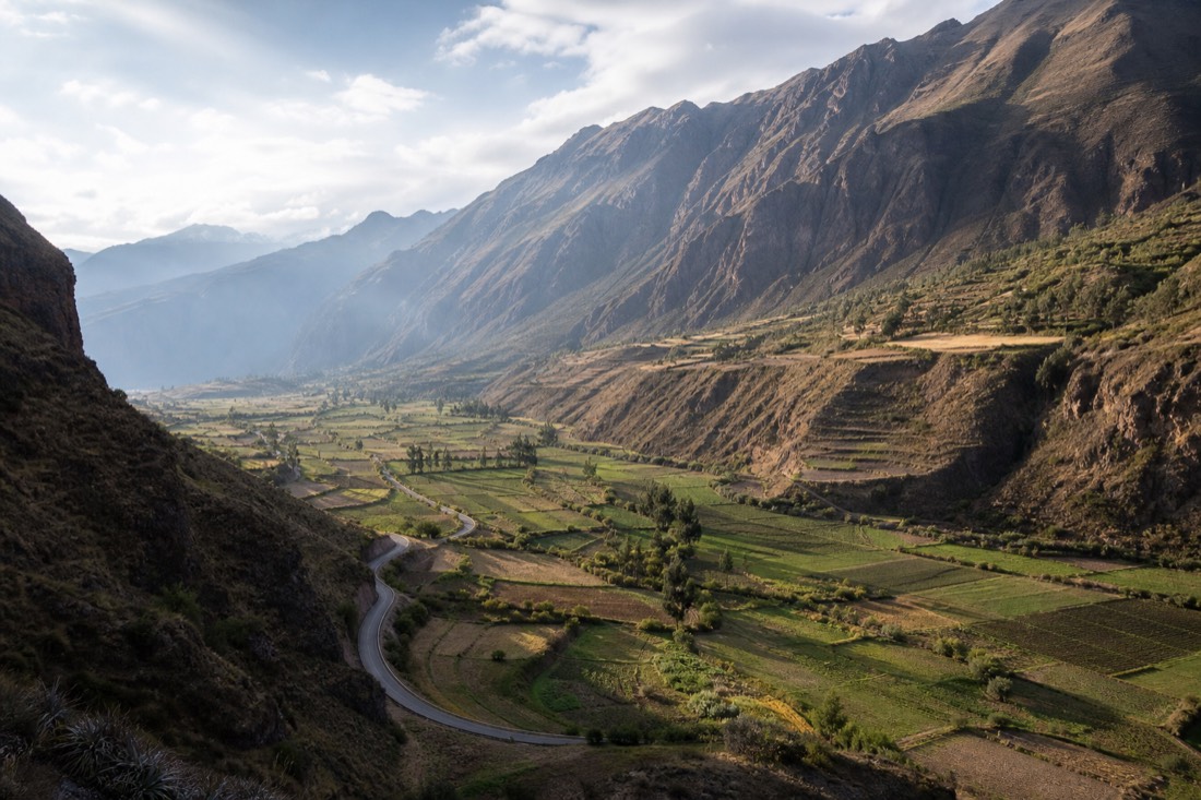 Afternoon light over green agricultural terraces in the Limatambo valley on the drive back to Cusco, a winding road and distant ridges, no people