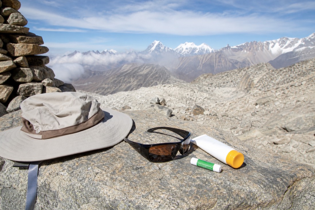 Sun protection kit on a rock at altitude: high-SPF sunscreen tube, wraparound sunglasses, a brimmed hat and lip balm, bright Andean daylight and pale scree, no faces, no logos