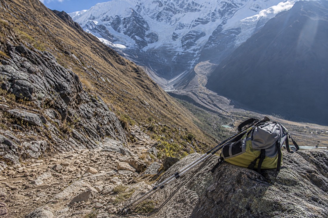 A lone hiker's daypack and trekking poles resting on a rock beside the steep rocky Humantay trail with the glacier valley ahead, no people